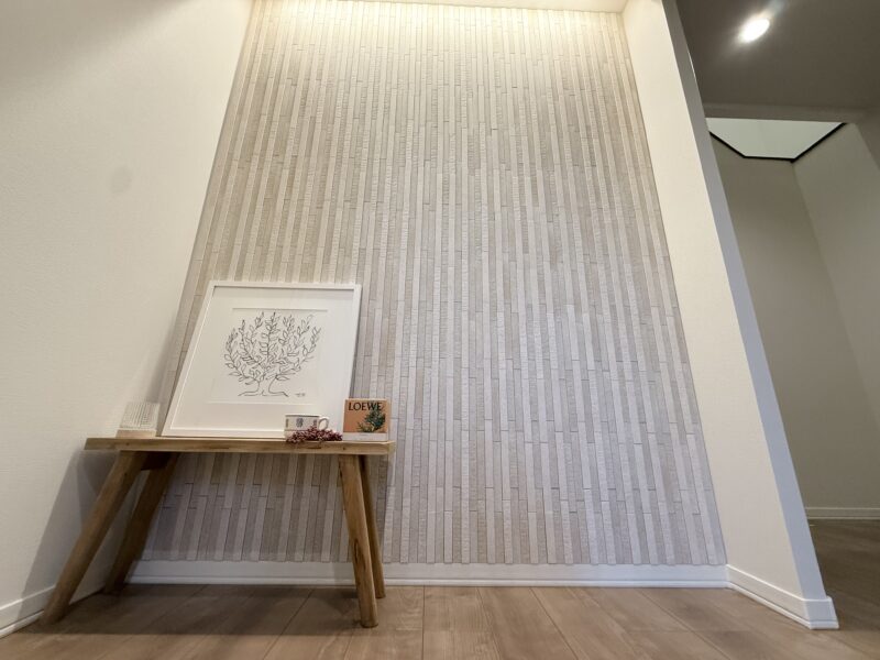 玄関ホール正面の壁一面にDIY施工したLIXILエコカラットプラス「陶連子」。天井からのコーニス照明が縦格子の凹凸を美しく照らし出し、陰影を作っている。手前には木製棚とアートフレームが飾られている。